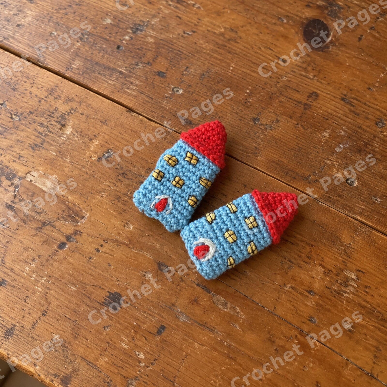 Two blue crochet house brooches with red roofs, embroidered doors and yellow windows.
