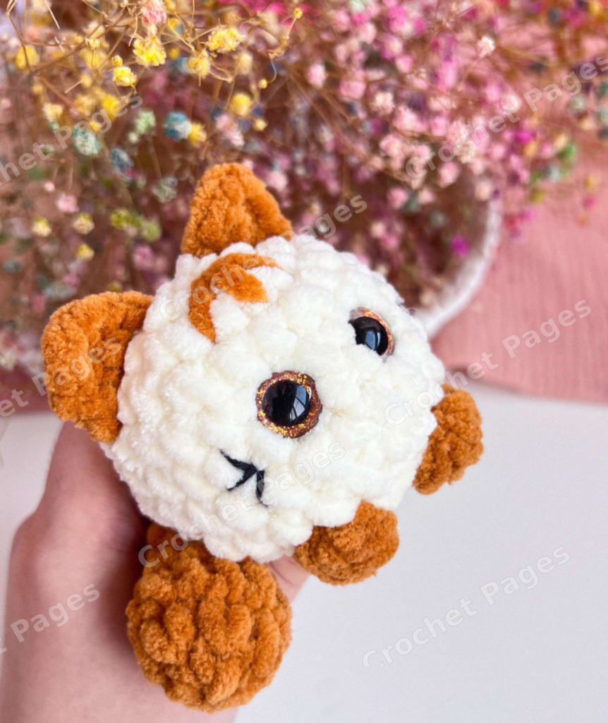 Make a small rounded plush cat with contrasting bobble paws, crocheted ears and a separate curled-looking tail. Finished-project view.