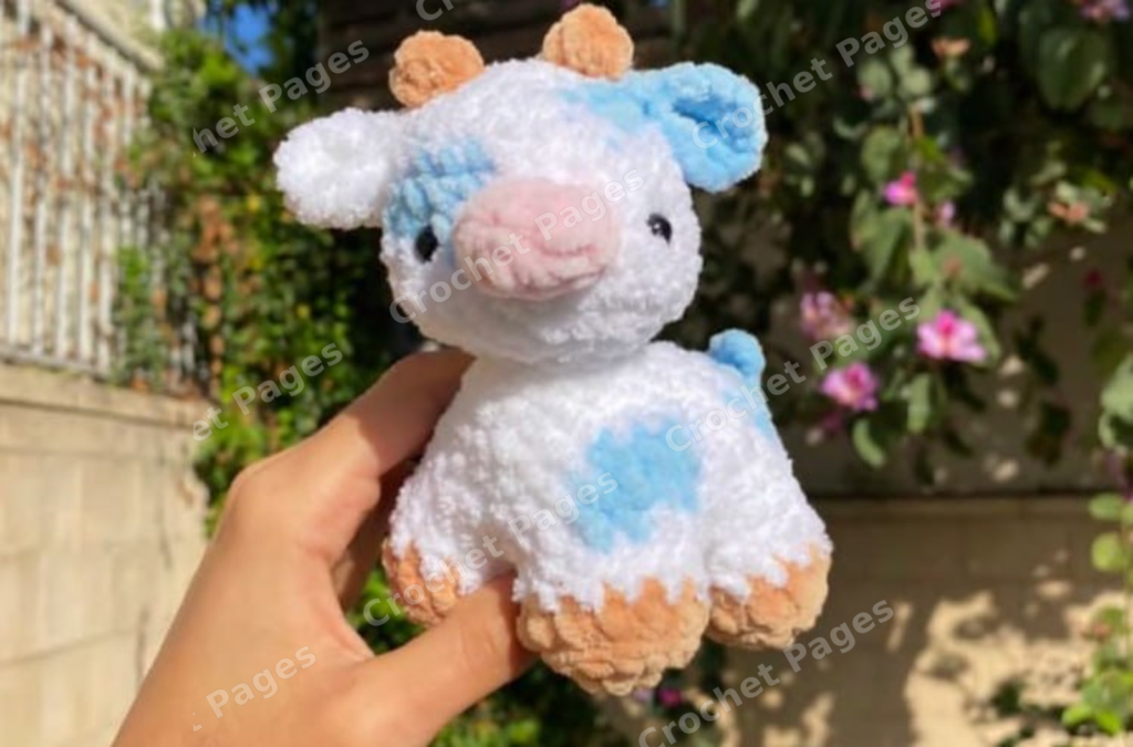 Make a four-legged cow with blue-and-white colorwork patches, tiny horns, folded ears, a pink muzzle and a short tail.